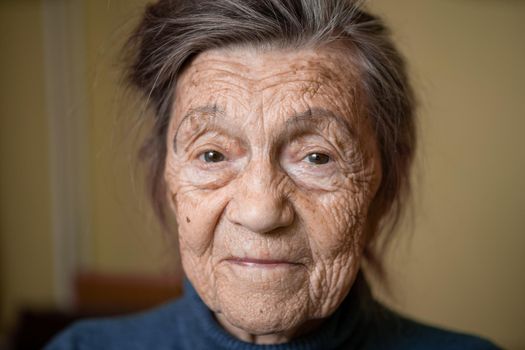 90 year old cute elderly woman with gray hair and wrinkles face, wearing sweater, portrait large, smiling and looking joyfully, background of room. Theme long-liver and aging, old people in good mood.