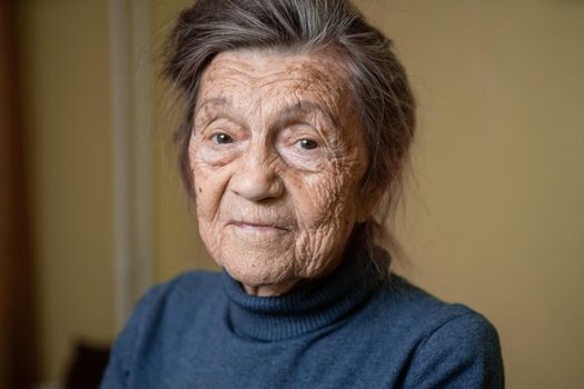 Older cute woman of ninety years old Caucasian with gray hair and wrinkled face looks at camera, cute kind look and smiles.Mature grandmother retired long-liver, theme emotion and mood of old people.