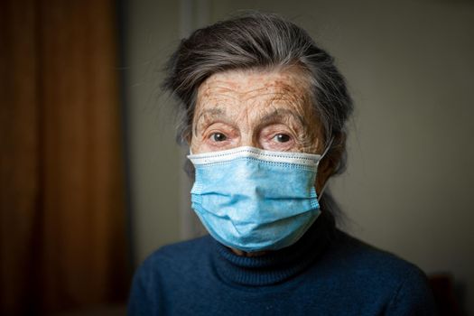 Elderly Caucasian woman with wrinkles on face, gray hair, wearing medical mask, looks attentively at camera, feeling need care and support. Topic safe communication, stay at home during diseases.