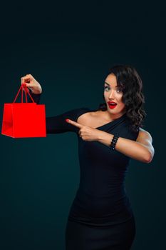 Beautiful young woman make shopping in black friday holiday. Girl with black bag on dark background. Beautiful girl with curly hair pointing to looking left.