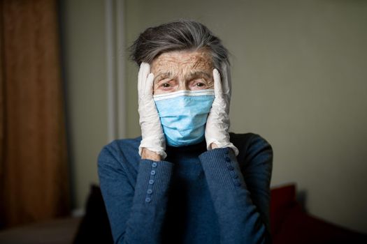 Sad senior woman in protective mask, latex gloves at home during coronavirus pandemic. Stay at home. Old granny retired quarantined epidemic covid 19. Gray hair female in mask infection, bacteria flu.