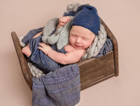 Little smiling baby weared in a blue knitted beanie sweetly sleeping in the small brown bed covered with blue-gray blankets ,on the light pink background