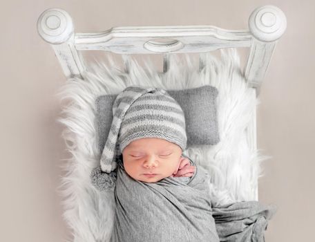 Cute little baby in hat sweetly sleeping in white bed covered with gray blanket