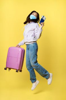 Full length shot happy asian woman jumping with suitcase and tickets on tour, holding passport, going on vacation, standing over yellow background.