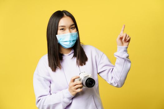 Happy asian girl in face mask, taking pictures, pointing finger at copy space, holding camera, standing over yellow background.