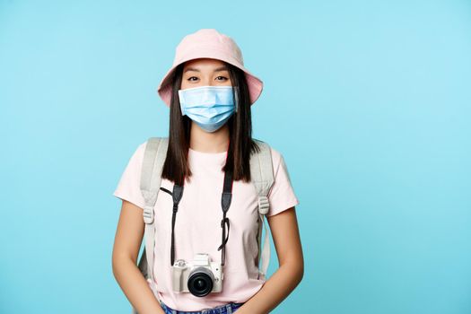 Smiling asian woman with medical face mask, travelling during pandemic, standing with photo camera, tourist abroad on vacation, blue background.