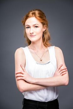 portrait of a young red-haired beautiful girl in the studio on a gray isolated background. A woman is standing with her arms folded and smiling in a white shirt with a short sleeve. Business concept