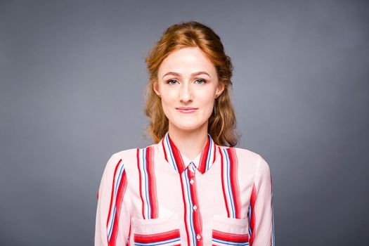 portrait of a young red-haired beautiful girl in the studio on a gray isolated background. A woman stands and smiles in a red shirt with a short sleeve. Business concept.