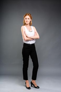 beautiful young Caucasian woman with long red hair in high heels, black trousers and a white shirt stands in full growth on a gray isolated background. Business concept