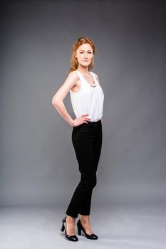 beautiful young Caucasian woman with long red hair in high heels, black trousers and a white shirt stands in full growth on a gray isolated background. Business concept