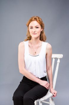 beautiful young caucasian woman with long red hair in high heels sitting on a chair, black trousers and white shirt in full growth on a gray background. Business concept.