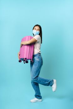 Full length of happy asian female tourist, dancing with suitcase in medical face mask, going on vacation, travelling with briefcase, standing over blue background.