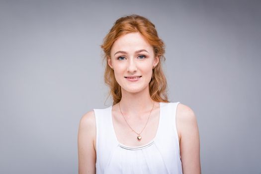 Closeup portrait young, beautiful business woman, student with lred, curly hair and freckles on face on gray background in the studio. Dressed in white blouse with short sleeves about open shoulders.