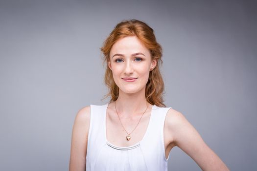 Closeup portrait young, beautiful business woman, student with lred, curly hair and freckles on face on gray background in the studio. Dressed in white blouse with short sleeves about open shoulders.