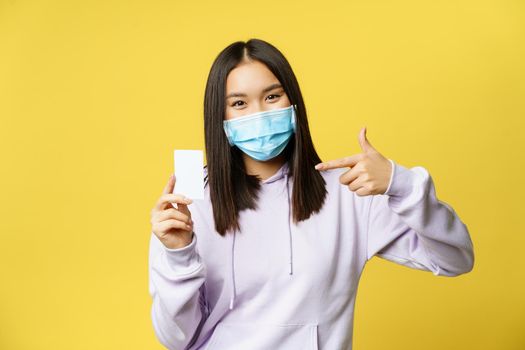 Cute korean woman in medical face mask, pointing at credit, discount card, showing smth, standing over yellow background.