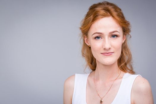 Closeup portrait young, beautiful business woman, student with lred, curly hair and freckles on face on gray background in the studio. Dressed in white blouse with short sleeves about open shoulders.