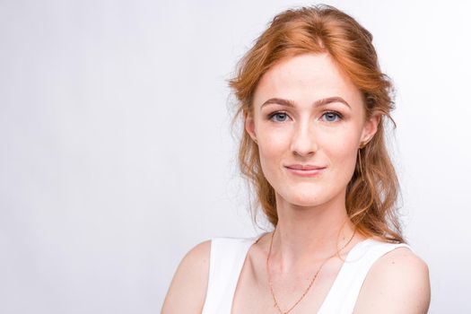 Portrait of a beautiful young woman of European, Caucasian nationality with long red hair and freckles on her face posing on a white background in the studio. Close-up student girl in a white blouse.