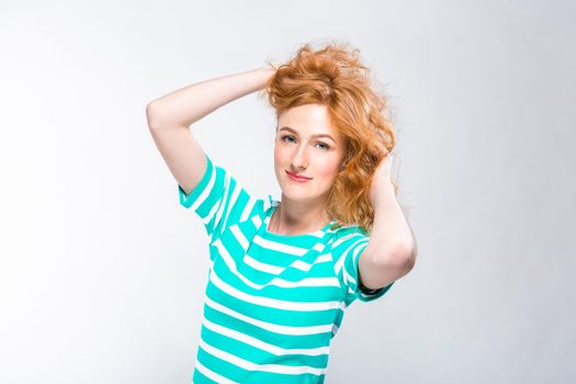 Close-up portrait of a young, beautiful woman with red curly hair in a summer dress with strips of blue in the studio on a gray background. Theme of summer vacation, tourism and summer clothes.