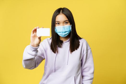 Smiling korean girl in face mask, showing card, standing in casual hoodie over yellow background.