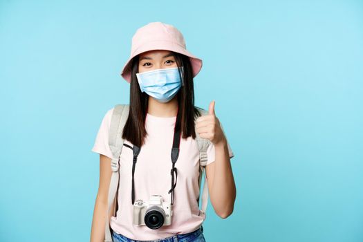 Cheerful asian woman in medical face mask, holding camera, showing thumbs up, travelling abroad, going on vacation. Tourist standing over blue background.