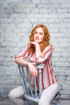 portrait Beautiful young woman student with red curly hair and freckles on her face sitting on a wooden chair on a brick wall background in gray. Dressed in a red striped shirt.