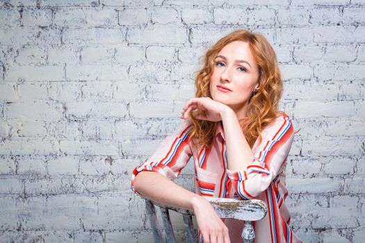 portrait Beautiful young woman student with red curly hair and freckles on her face sitting on a wooden chair on a brick wall background in gray. Dressed in a red striped shirt.