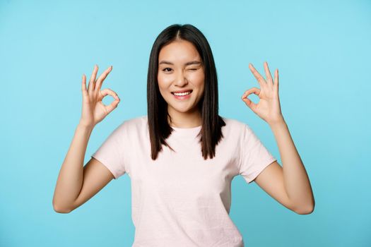 Smiling korean woman winking, showing okay signs, recommending company or store, standing in t-shirt over blue background.