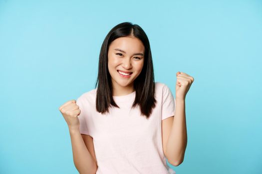 Enthusiastic cute asian girl winning, rejoicing from great news, victory dance, smiling pleased, standing over blue background.