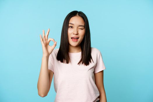 Satisfied asian girl winks and shows ok sign, give approval, recommends, stands in t-shirt over blue background.