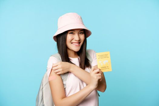 Smiling korean tourist, happy woman shows her arm vaccinated arm and covid-19 international vaccination certificate, standing over blue background.