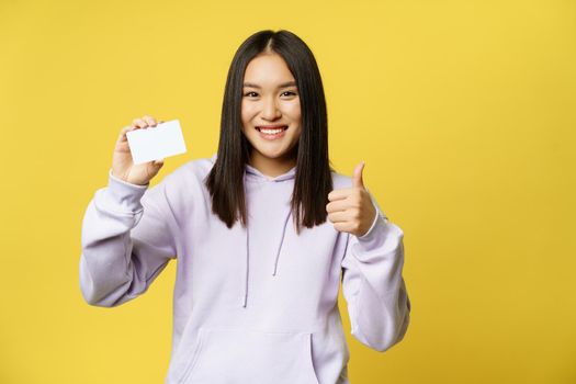 Smiling japanese girl showing card and looking pleased, thumbs up gesture, standing over yellow background.