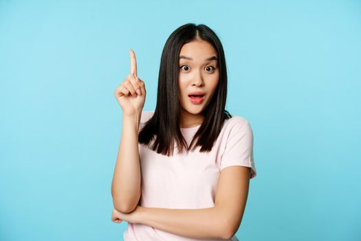 Creative korean girl raising finger up, eureka gesture, showing advertisement, looking surprised, standing over blue background.
