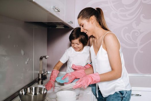 housewife mom in pink gloves washes dishes with her son by hand in the sink with detergent. A girl in white and a child with a cast cleans the house and washes dishes in homemade pink gloves.A child with a cast washes dishes and smiles.