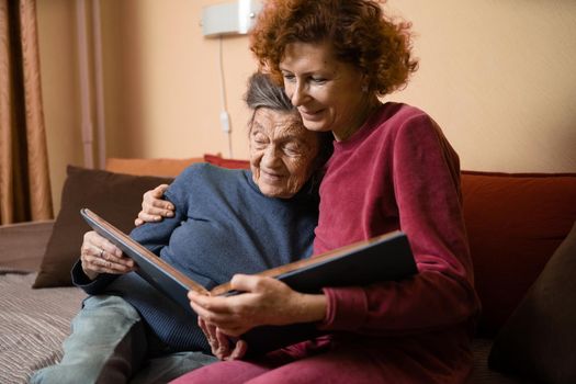 Mature daughter visits senior mother and has fun watching family photo album, sitting in embrace in living room couch. Mothers Day. Elderly woman and old gray hair grandmother laugh and remember youth