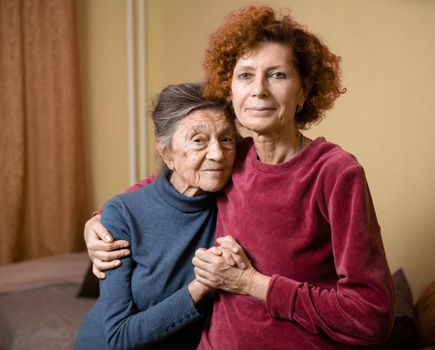 Senior Caucasian woman tenderly hugs her ninety year old elderly mother with gray hair, face with deep wrinkles, at home on sofa, smiling mother and daughter. Theme care and dedication to old parents.