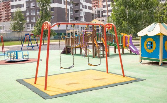 A colorful modern playground in the courtyard in a park without people. Swing, outdoor carousel for children. A playground in a public park surrounded by green trees and new houses. Children exercise