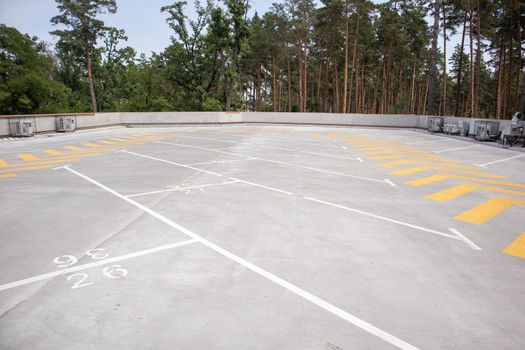 Empty rooftop parking. Directional traffic arrows and parking spots are marked with white and yellow paint on the asphalt. Parking space without cars with city skyline and park with trees