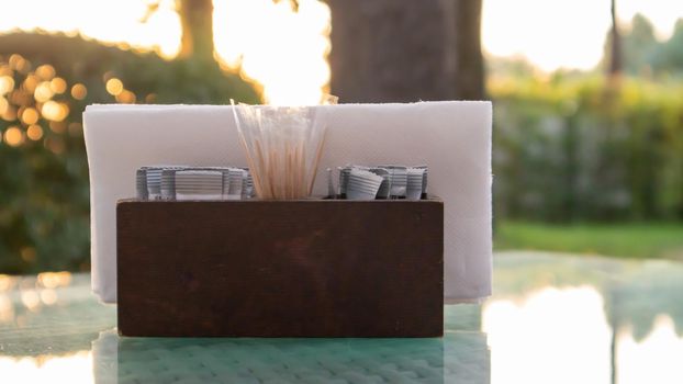 A brown wooden napkin holder and toothpicks sit on the glass table of the cafe, illuminated by the sun's rays. Table setting in the restaurant. Glass table in a green outdoor garden
