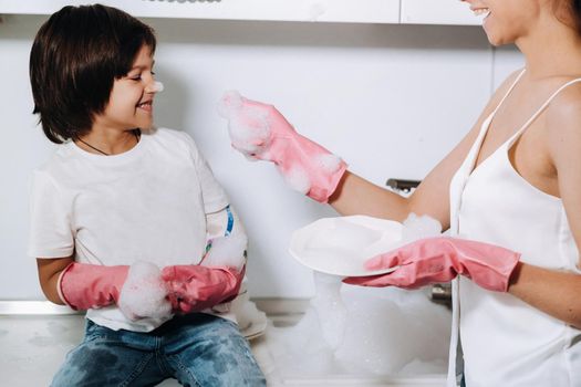 housewife mom in pink gloves washes dishes with her son by hand in the sink with detergent. A girl in white and a child with a cast cleans the house and washes dishes in homemade pink gloves.A child with a cast washes dishes and smiles.