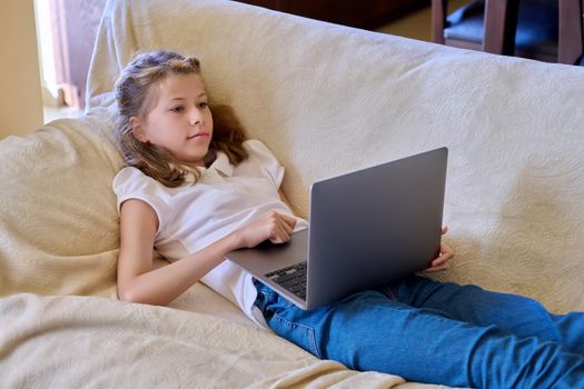 Child girl with laptop at home on the couch. Preteen blonde girl looking at laptop screen, education, relaxation, leisure, computer game, video communication, home lifestyle