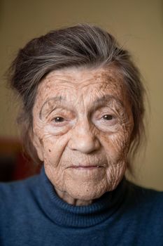 90 year old cute elderly woman with gray hair and wrinkles face, wearing sweater, portrait large, smiling and looking joyfully, background of room. Theme long-liver and aging, old people in good mood.