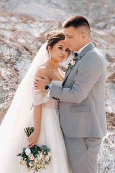 A beautiful couple of lovers posing in a white salt mountain. A young woman in a stylish wedding dress and a beautiful stylish man in a gray suit. The concept of the wedding day