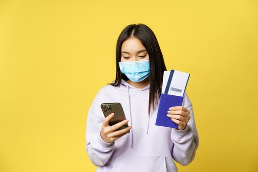 Japanese girl in face mask, holding her passport and flight tickets, using smartphone app, going on vacation, travelling during covid pandemic, yellow background.