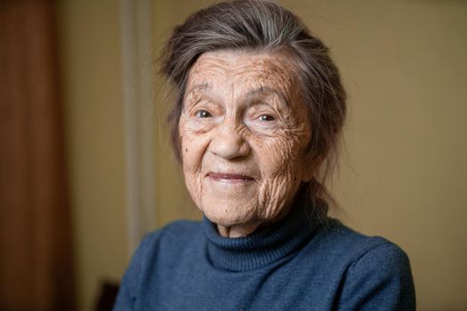 90 year old cute elderly woman with gray hair and wrinkles face, wearing sweater, portrait large, smiling and looking joyfully, background of room. Theme long-liver and aging, old people in good mood.