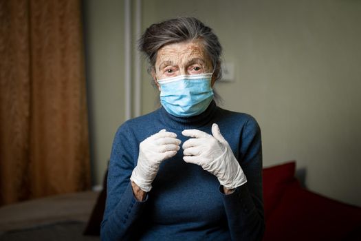 Senior woman in protective medical mask and latex gloves stands and watches into camera with sad, tired, upset emotions at home. Theme lockdown, stay home, coronavirus and covid 19.