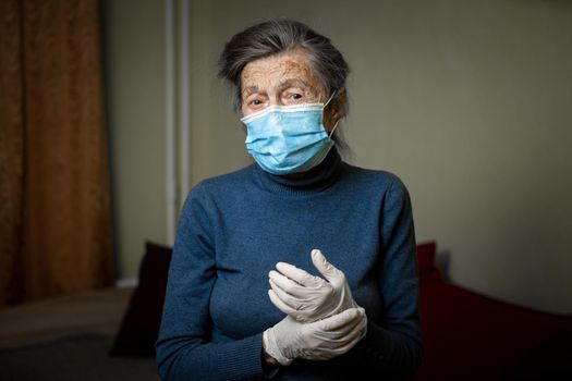 Old Caucasian woman dressed in personal protective equipment, mask and gloves, encourages to stay at home during an epidemic for safety. Health care and elderly care theme.