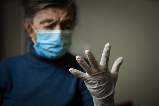 White medical glove in foreground, shown by an elderly grandmother wearing a medical mask, calling for safety and sanitation during an epidemic. Senior woman in personal protective equipment.