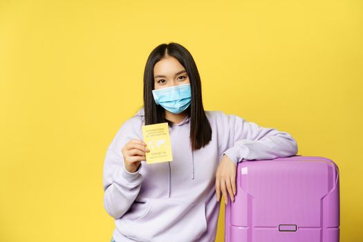 Covid-19 and healthcare concept. Tourism. Young asian woman tourist shows her health passport, coronavirus international vaccination certificate, standing with suticase.