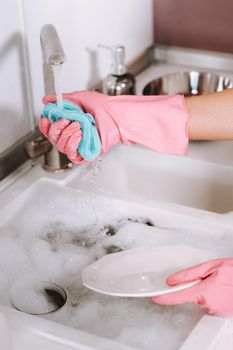housewife girl in pink gloves washes dishes by hand in the sink with detergent. The girl cleans the house and washes dishes in gloves at home