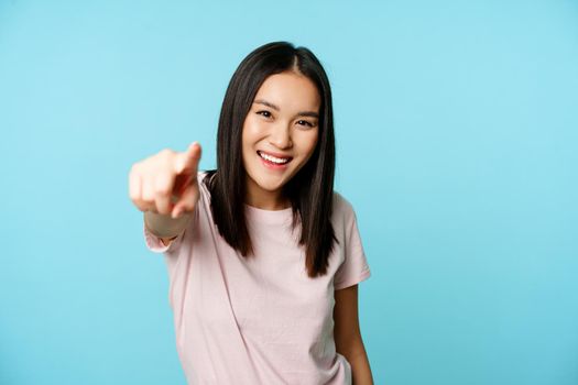 Its you. Smiling happy asian woman pointing finger at camera, congratulating, inviting people, standing in t-shirt over blue background.